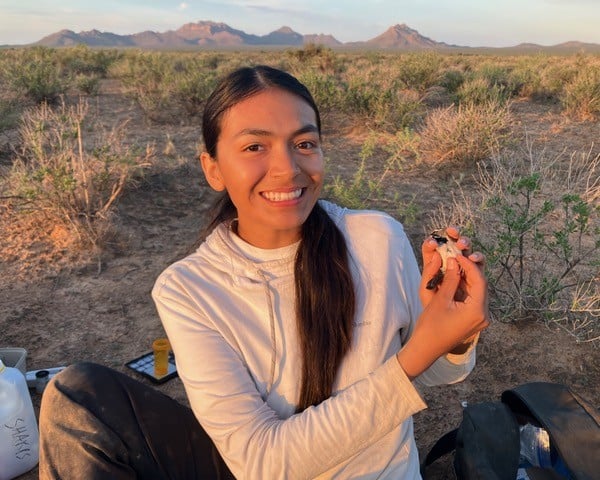 Amyliah Pina in the desert holding a small bird in her hands. A low sun emits golden light and casts shadows on the mountains behind her.