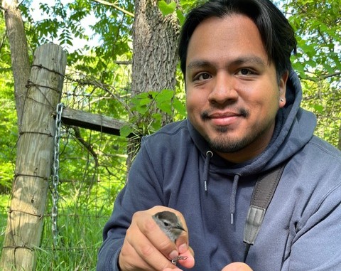 Eriberto Osorio smiling and holding a small gray bird in his hand. He is wearing a navy sweatshirt.