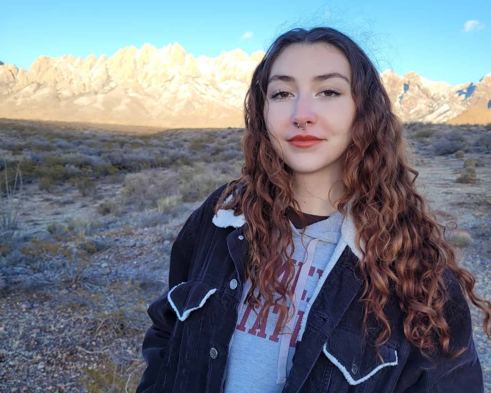 Julianna Diaz standing before the snow-covered Organ Mountains. She has brown curly hair and is wearing a black jacket over a gray New Mexico State sweatshirt.