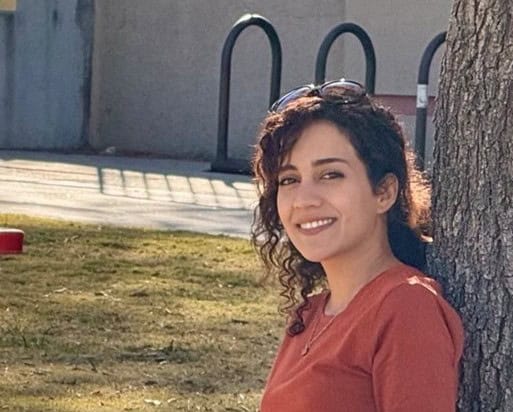 Negin Jazi smiling and leaning against a tree. She is wearing an orange top.