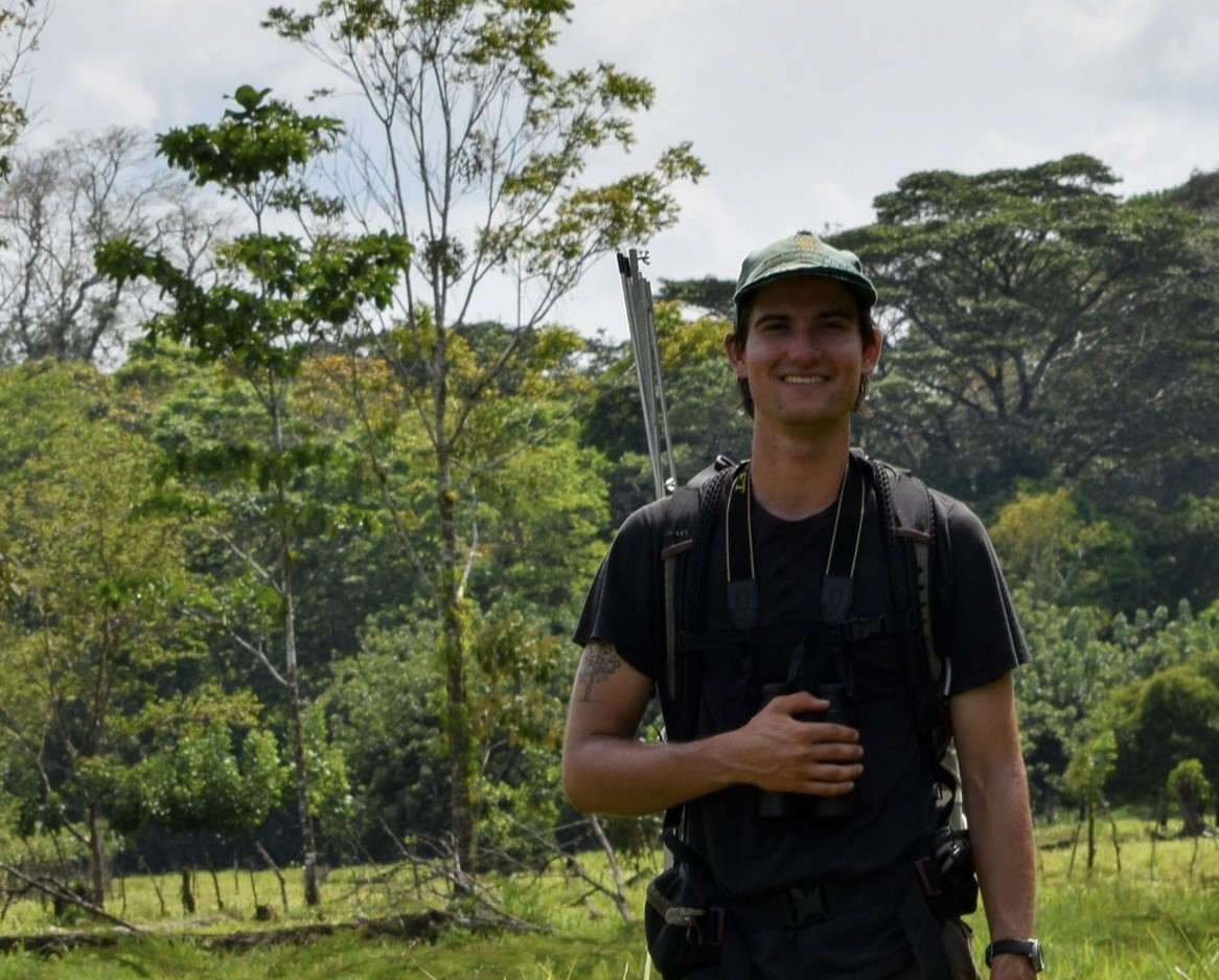 Alexander Allison smiling while holding binoculars to his chest. The background is full of tall, green trees.