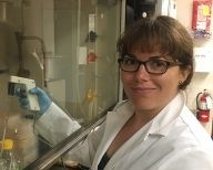 Amelia Clayshulte Abraham pipetting in a hood. She is smiling at the camera and is wearing a white lab coat and light blue nitrile gloves.