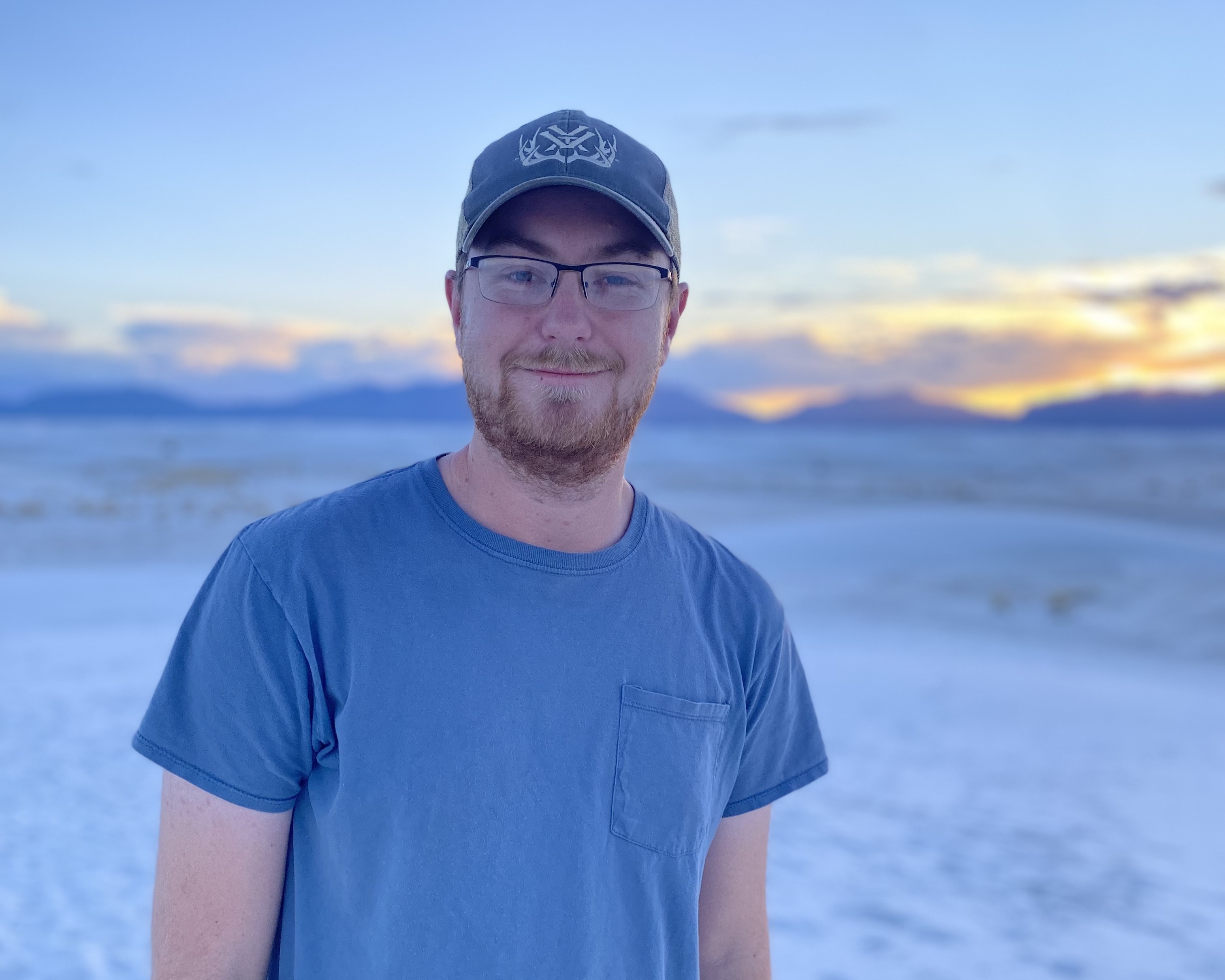 Dylan Osterhaus is wearing a dark sky blue shirt and a matching baseball cap. White Sands National Park at sundown is the background.