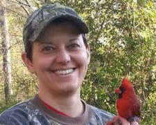 Jodie Jawor smiling at the camera with a cardinal held by its legs in her hand. She is wearing a camo shirt and a camo baseball cap.