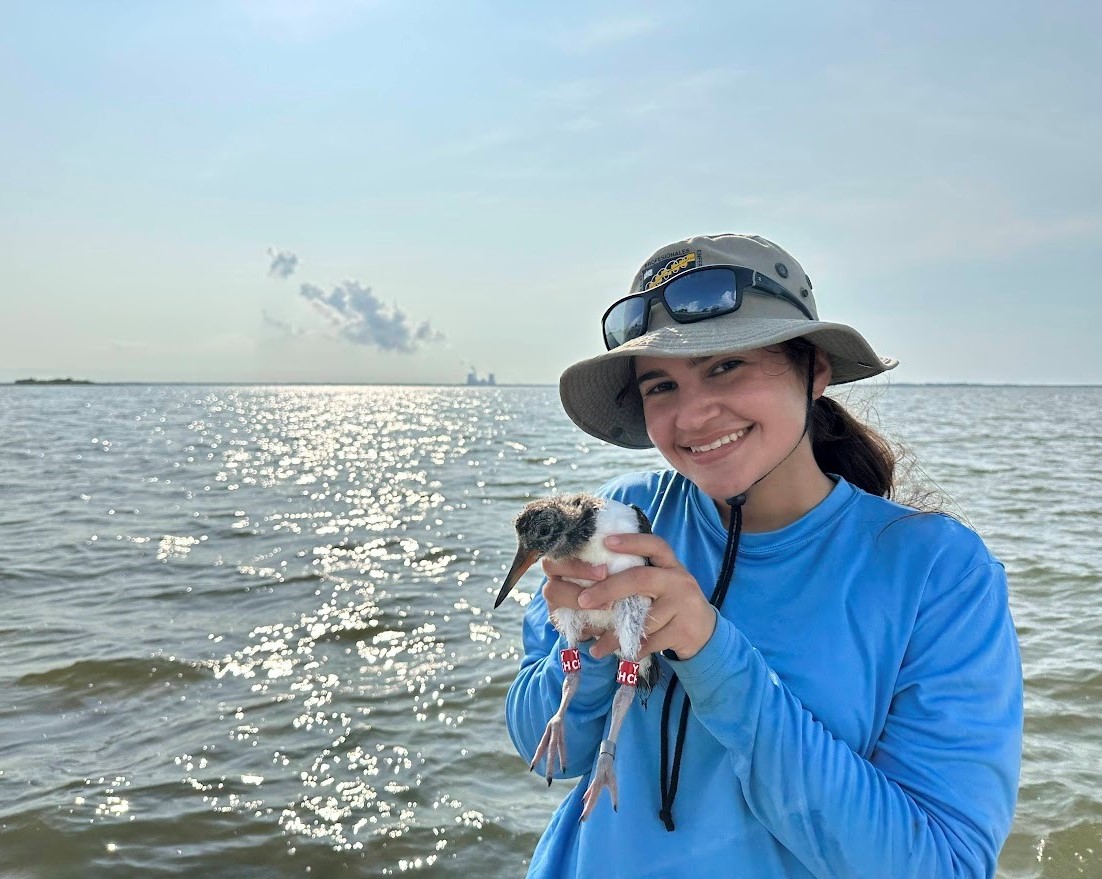 Juliemar is standing before a body of water holding a type of water bird with both hands. She is smiling and is wearing a bright blue shirt and a light brown bucket hat with black sunglasses on top.