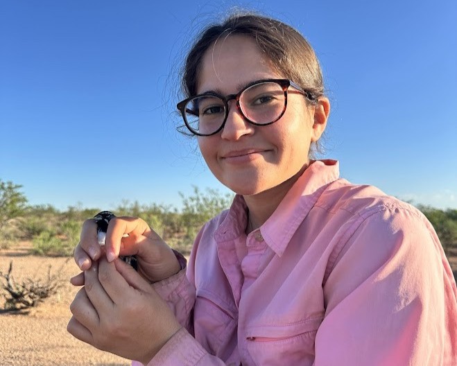 Juliemar crouches in the desert holding a bird in a pink shirt and black-framed glasses.