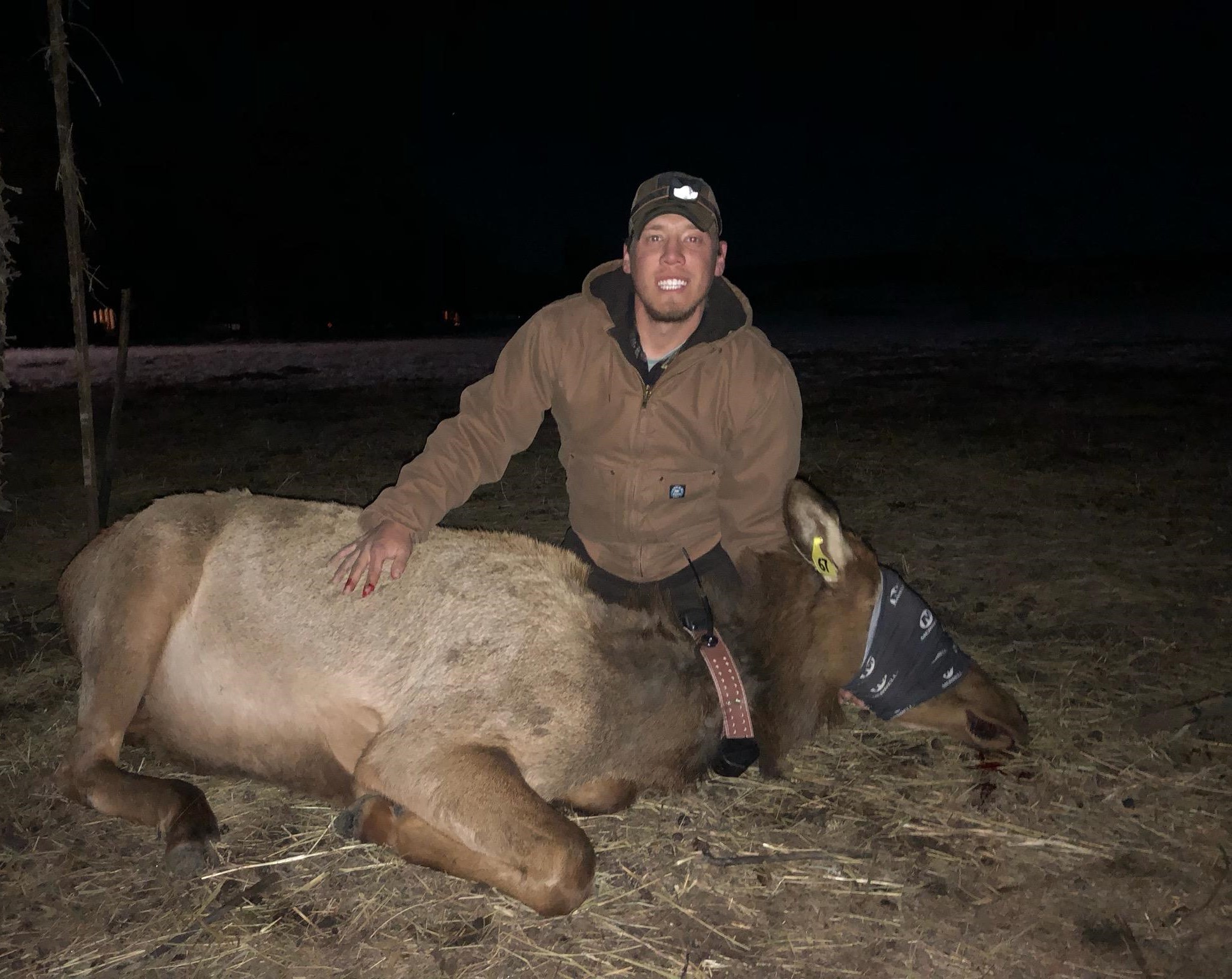 Scott Boyle smiling & wearing a dark brown heavy jacket and brown baseball cap. He is in front of a deer that has its eyes covered.