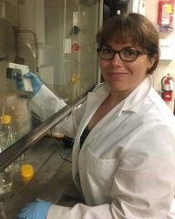Amelia Clayshulte Abraham pipetting in a hood. She is smiling at the camera and is wearing a white lab coat and light blue nitrile gloves.